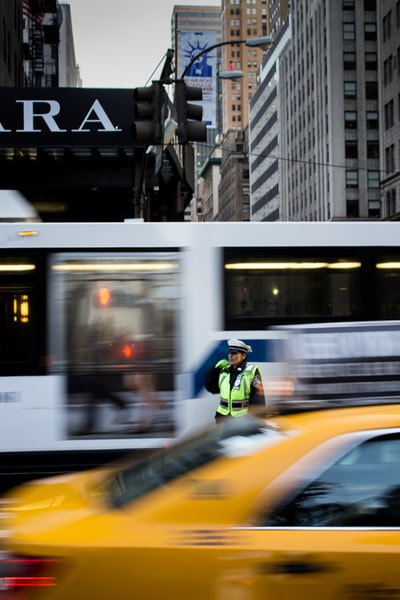身穿绿色安全背心和白色山顶帽的男子站在白色公共汽车和橙色汽车之间的照片